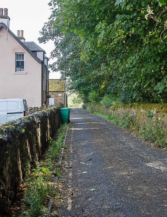 Woolmill Road - a narrow road bordered by low stone walls runs between residential buildings and green trees.