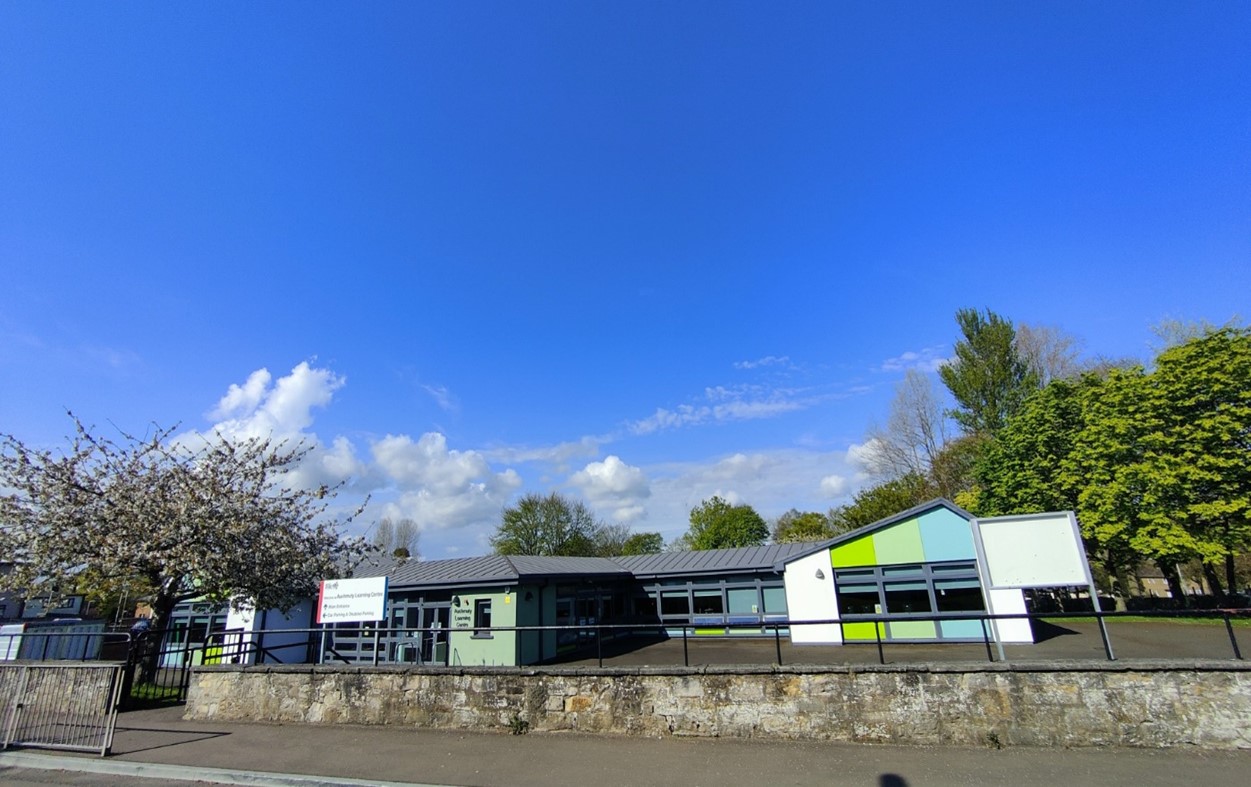 A brightly lit, wide-angle shot of a modern community building with colorful green, blue, and white paneling. The single-story building is set back from a stone wall, with lush green trees and a clear blue sky overhead.