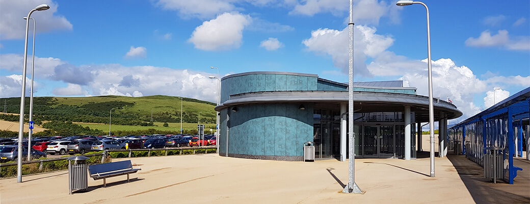 Halbeath Park & Ride waiting area with a curved modern building, large glass windows, nearby bench and bin, street lamps, and a car park in the background. Green hills and a blue sky with clouds are visible behind.